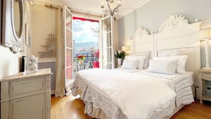 A bedroom with a large bed, a dresser, a nightstand, a chandelier, and a balcony with a view of the city. - One-of-a-Kind 3-bedroom OMG Eiffel Views & Wraparound Terrace (Paris)