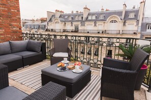 Ein Balkon mit Korbmöbeln, einem Tisch mit Croissants und Obst, und Blick auf historische Gebäude.