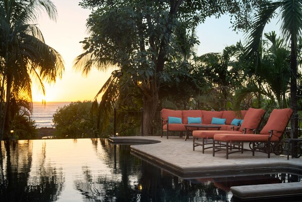 An infinity pool with a sunset view, surrounded by palm trees and outdoor furniture.