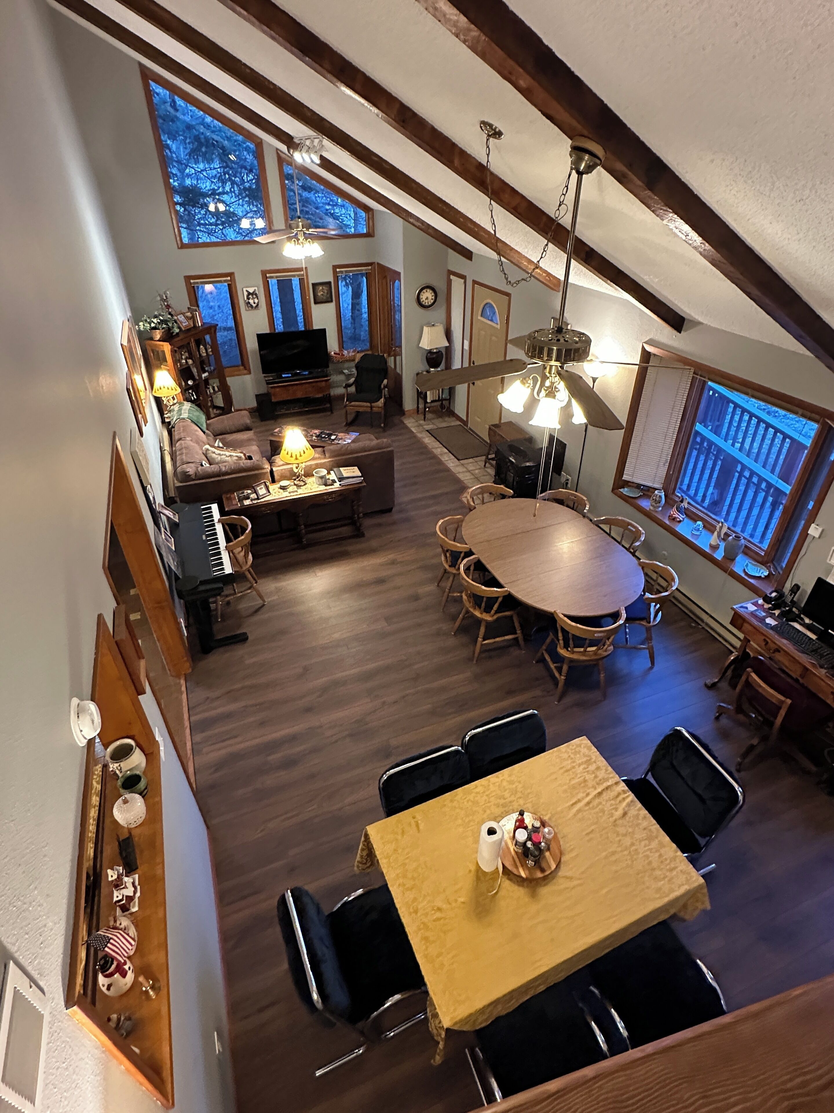 A spacious loft with a high ceiling, wooden beams, a dining area with a round table, a living room with a sofa and TV, and large windows offering views.