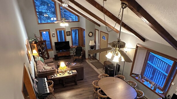 Um loft espaçoso com pé-alto, vigas de madeira, área de jantar com mesa redonda, sala de estar com sofá e televisão, e grandes janelas com vista.