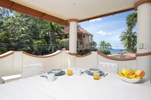 A balcony with a table set for breakfast, overlooking a tropical landscape and a building with a red roof. - 4 BR/4 BA, Pool, Ocean View, Steps from the Beach, Reduced Rates (Playa Potrero)