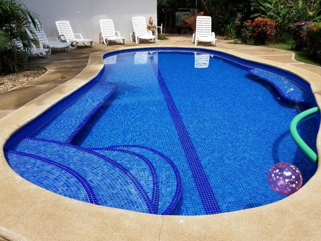 A swimming pool with a tiled edge, surrounded by white lounge chairs and a garden with plants and flowers.