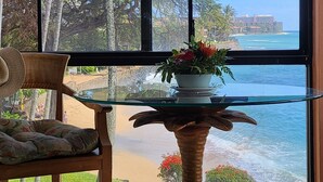 Una habitación con un ventanal grande con vistas a una playa y al océano, una mesita de vidrio con una planta en maceta y una silla de madera.