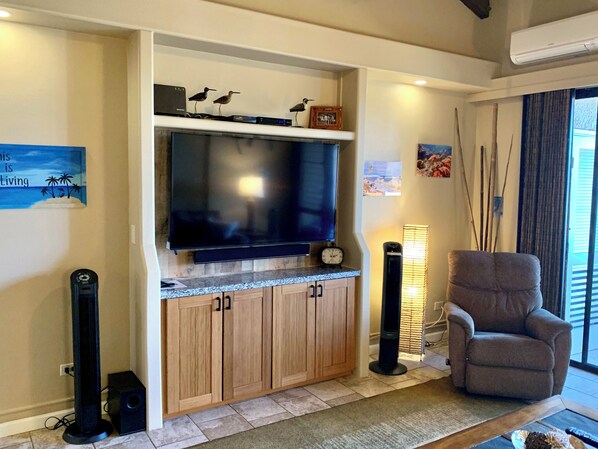 A living room with a flat-screen TV, wooden cabinets, a comfortable armchair, a standing speaker, and a wall-mounted clock.