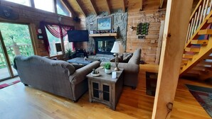 A cozy living room with a stone fireplace, wooden furniture, and a view through large windows.
