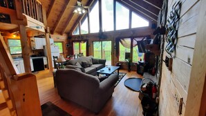 A spacious cabin with a living area featuring a sofa and a coffee table, a kitchen with white cabinets, and large windows offering views of the outdoors.