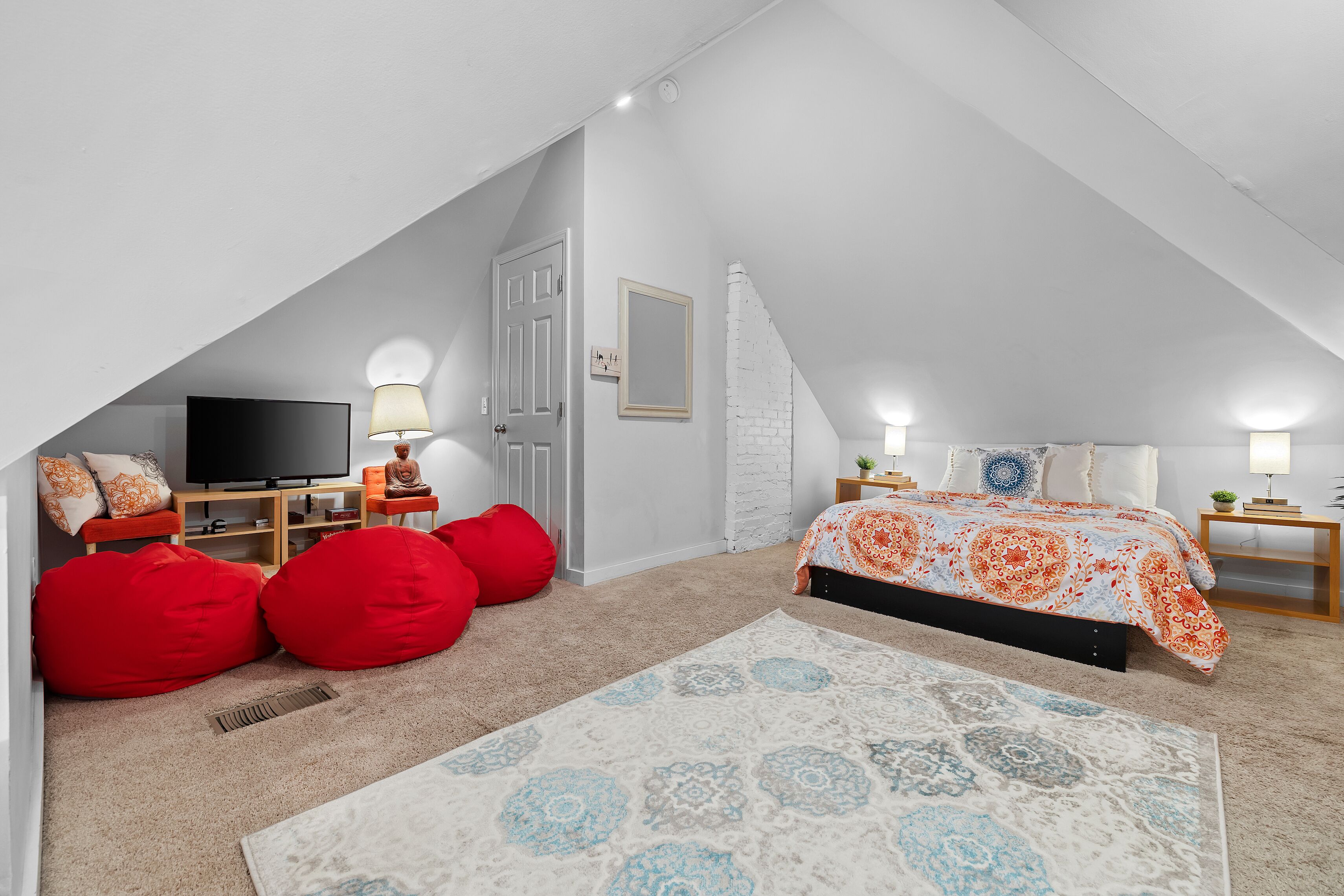 Une chambre avec un plafond en pente, un lit recouvert d’une couvre-lit à motifs orange et blanc, un téléviseur, un fauteuil en forme de poire rouge et une petite table de chevet avec une lampe.