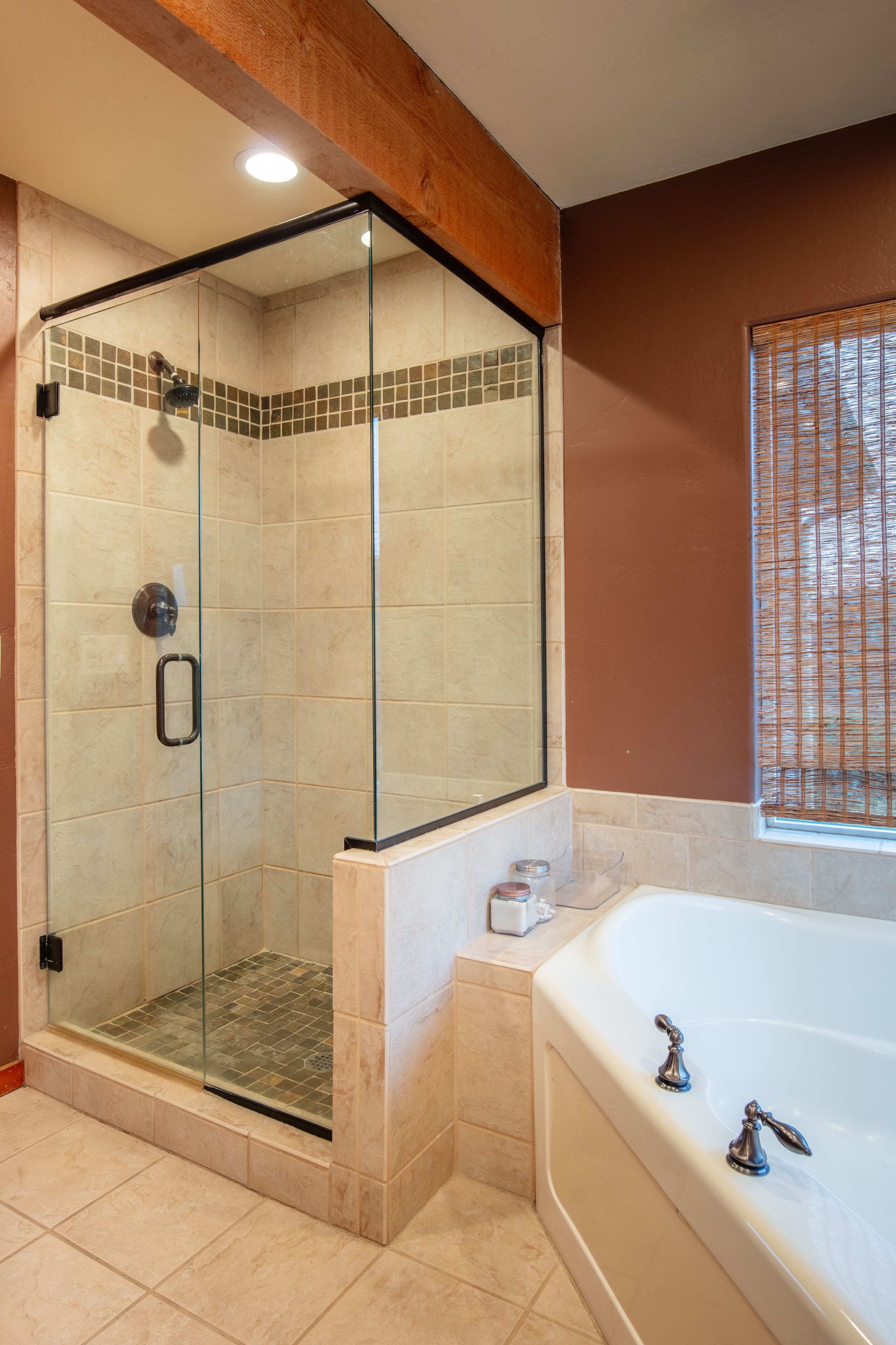 A modern bathroom with a glass-enclosed shower, a large bathtub, and a window with blinds.