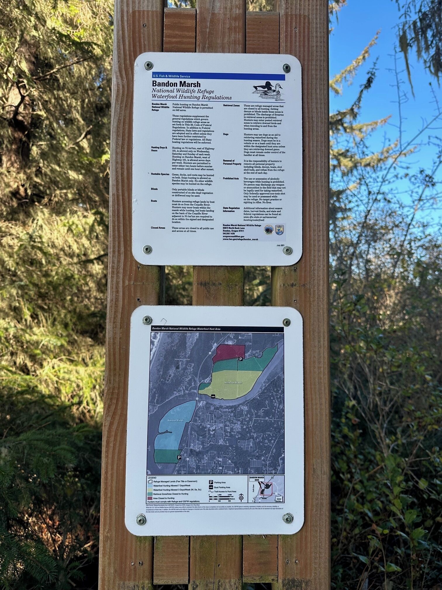 A wooden sign with two informational panels. The top panel details the Bandon Marsh National Wildlife Refuge's waterfowl hunting regulations, including hunting hours, prohibited acts, and a map of the refuge. The bottom panel features a color-coded map indicating different zones within the refuge.