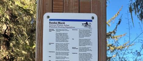 A wooden sign with two informational panels. The top panel details the Bandon Marsh National Wildlife Refuge's waterfowl hunting regulations, including hunting hours, prohibited acts, and a map of the refuge. The bottom panel features a color-coded map indicating different zones within the refuge.