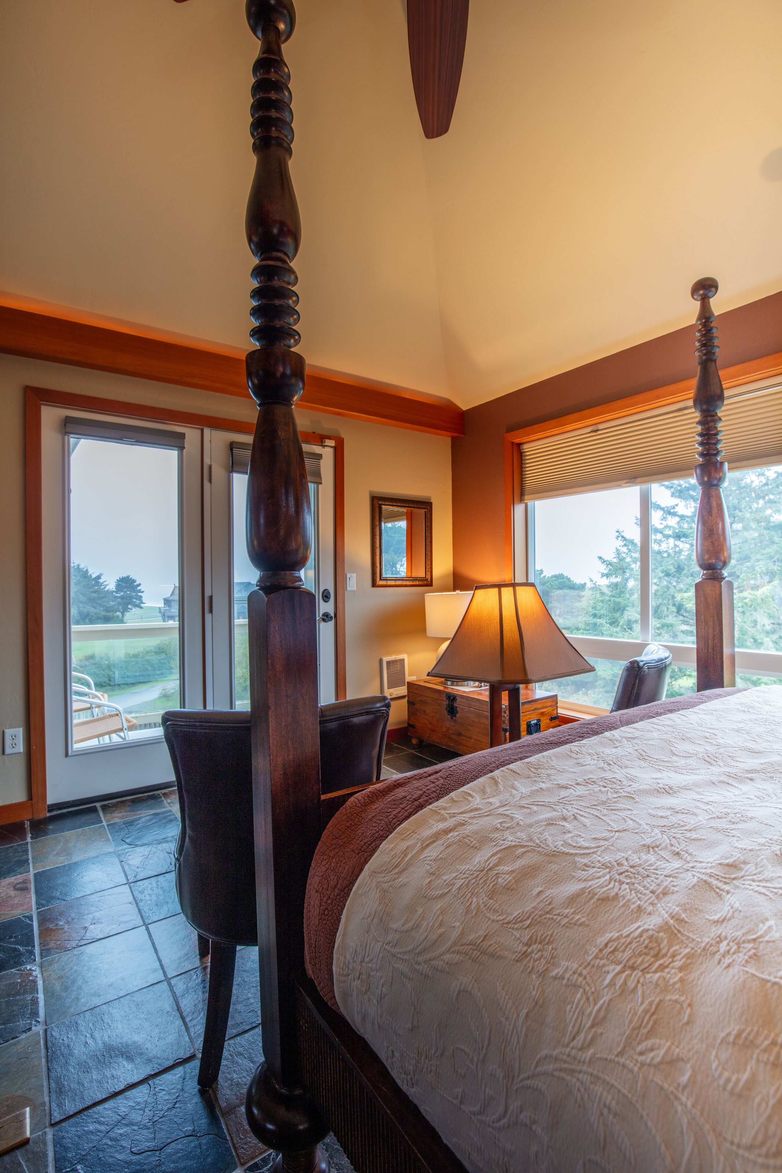 A bedroom with a four-poster bed, a desk with a lamp, and a view of the outdoors through a sliding door.