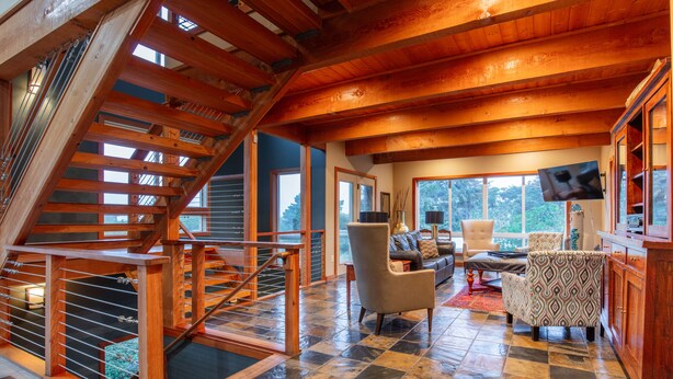 A spacious living area with a wooden staircase, comfortable seating, and large windows offering a view of the outdoors. -  Beach House W Ocean Views, 3 Golfer en suites, hot tub, fireplace, Pets OK! (Bandon)