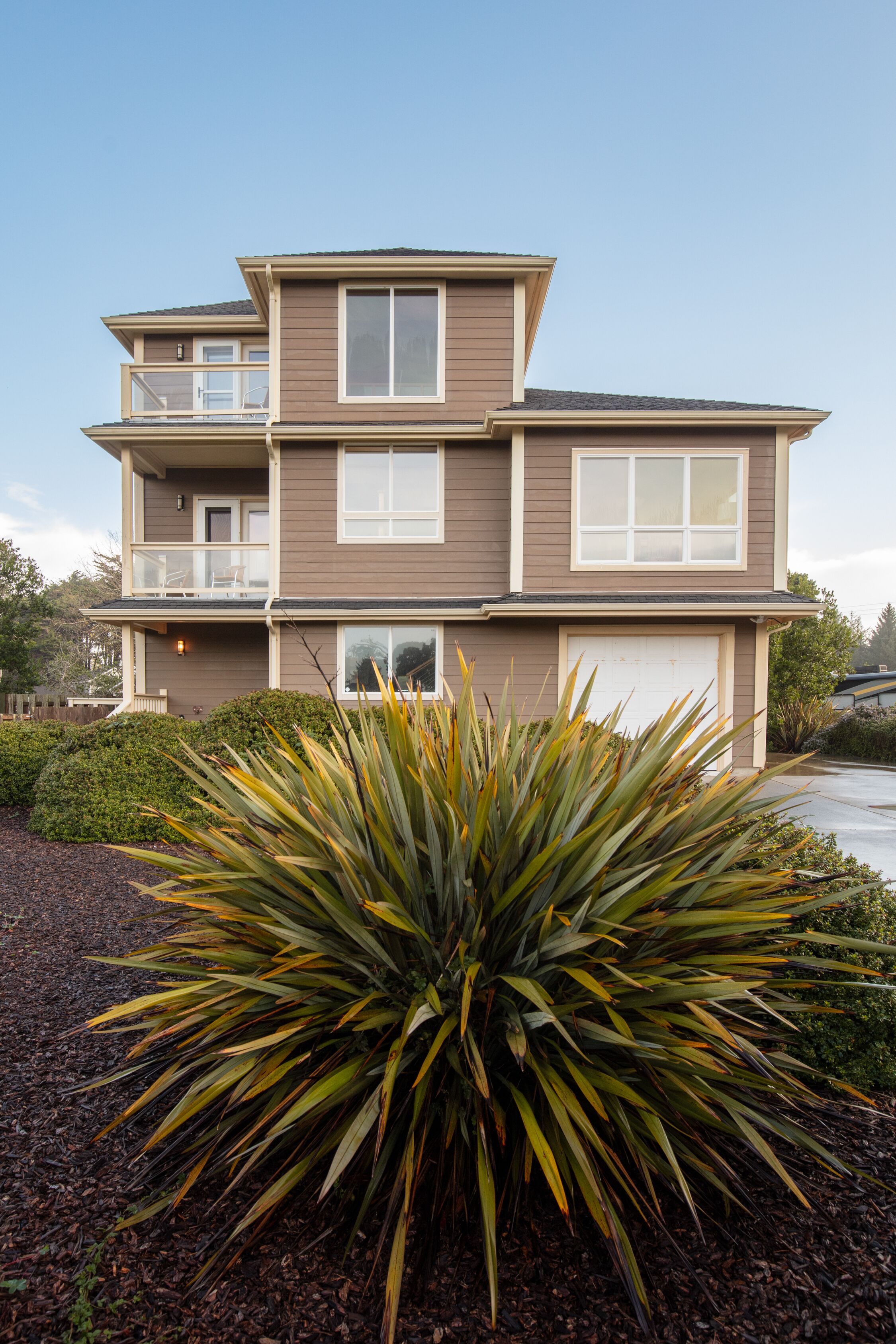 Une maison à deux étages avec un balcon à l’étage supérieur, un garage et un jardin bien entretenu agrémenté d’une grande plante d’agave.