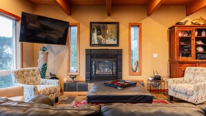 A cozy living room with a fireplace, patterned chairs, a large flat-screen TV, and a wooden cabinet with decorative items. -  Beach House W Ocean Views, 3 Golfer en suites, hot tub, fireplace, Pets OK! (Bandon)