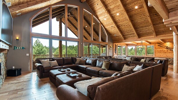 A spacious living area with a large L-shaped sectional sofa, wooden flooring, and a high ceiling with exposed beams.