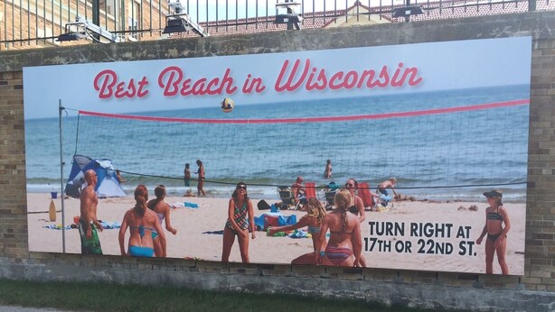 Strandschild, das 'Bester Strand in Wisconsin' bewirbt, mit einem Volleyballnetz und Personen am Strand.