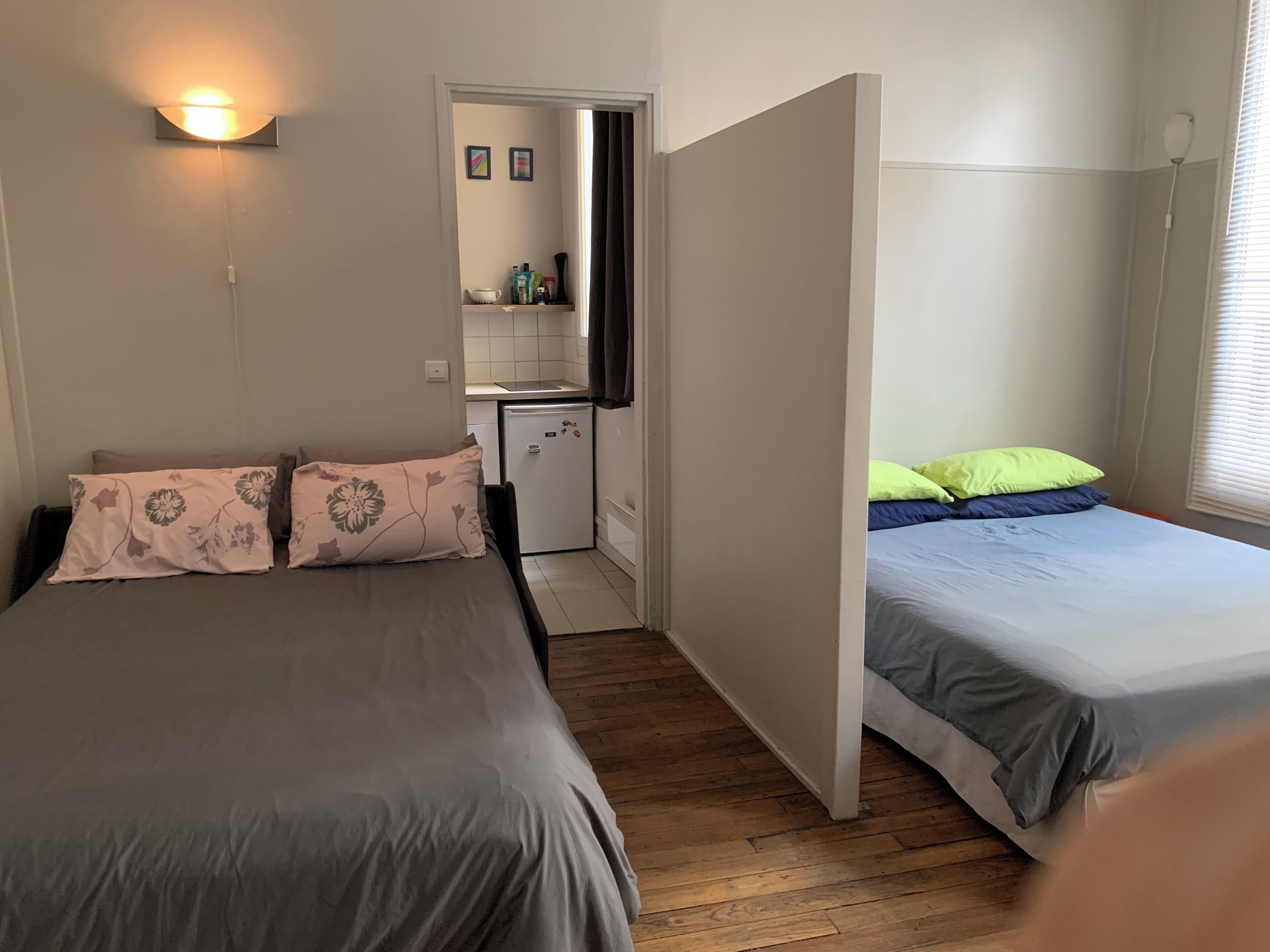 A bedroom with two beds, a small kitchenette, and a bathroom visible through a partition.