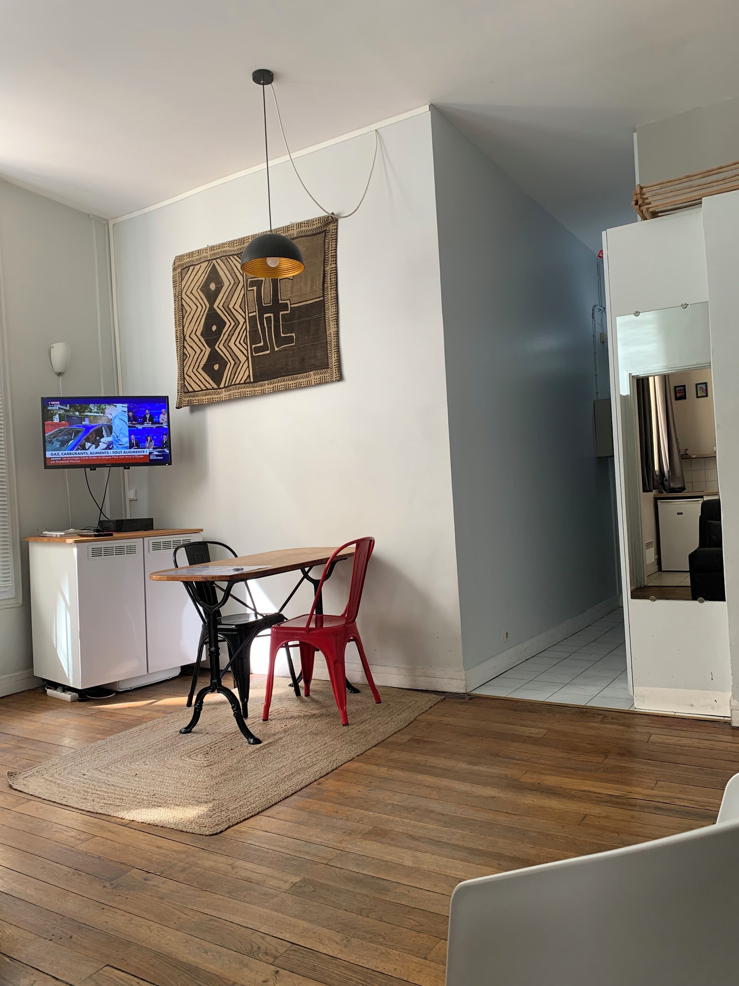 A compact living space with a kitchenette, a dining area with a table and chairs, a television mounted on the wall, and a patterned artwork above the table.