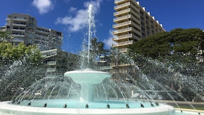 Une fontaine dotée de plusieurs jets d’eau, entourée d’un bassin circulaire. Au fond, on aperçoit des immeubles à plusieurs étages, dotés de balcons.