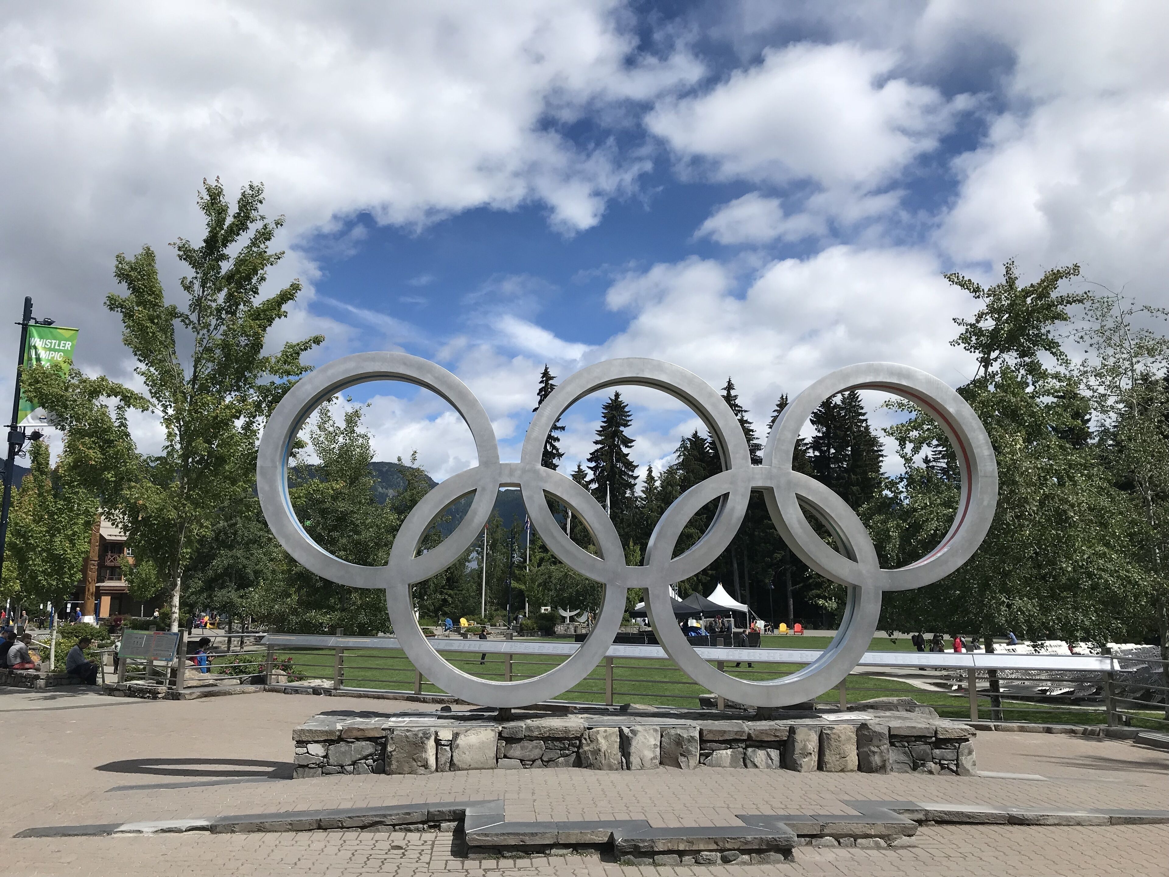 Een groot beeld van de Olympische ringen in een park met stenen paden en bomen.