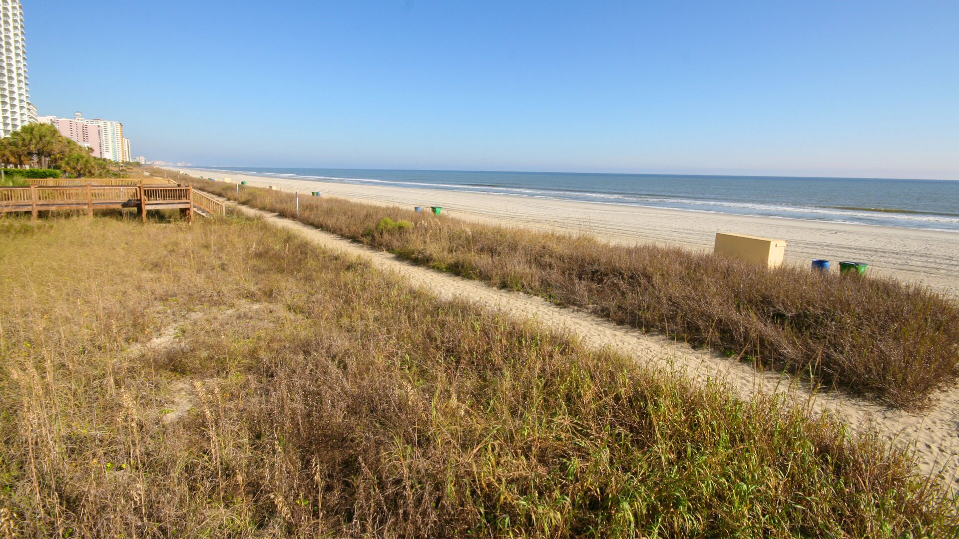A beach path with a wooden boardwalk leading to the shore, a sandy beach, and a clear blue sky.
