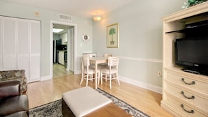 Un salon avec un canapé, une table à manger entourée de chaises, un téléviseur posé sur un meuble et, au fond, une cuisine.