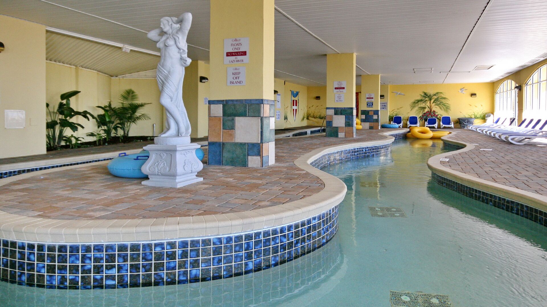 An indoor swimming pool with a central statue, tiled walls, and lounge chairs along the sides.