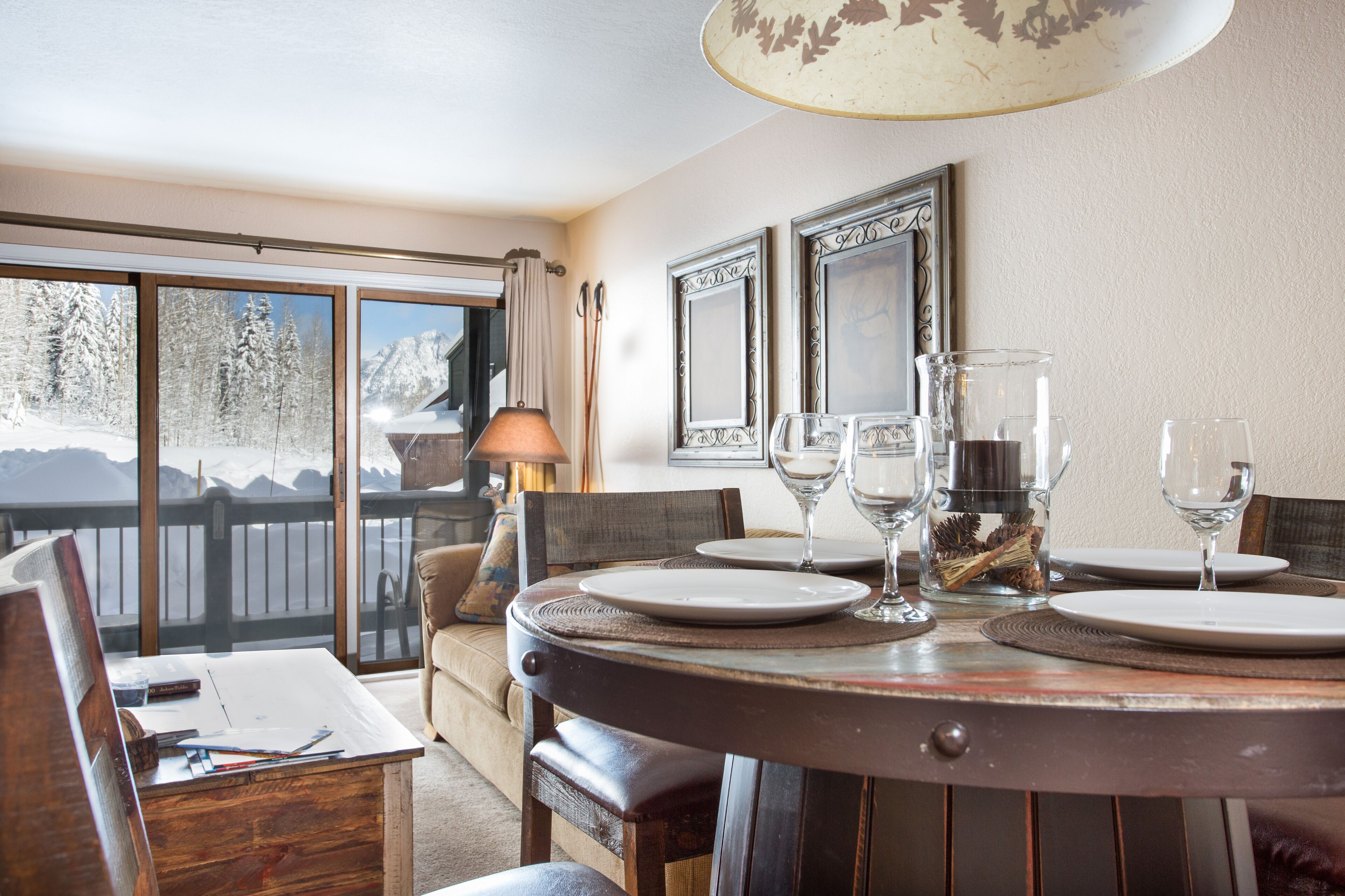 A dining area with a wooden table set for four, large windows with a snowy mountain view, and a lamp on the table.