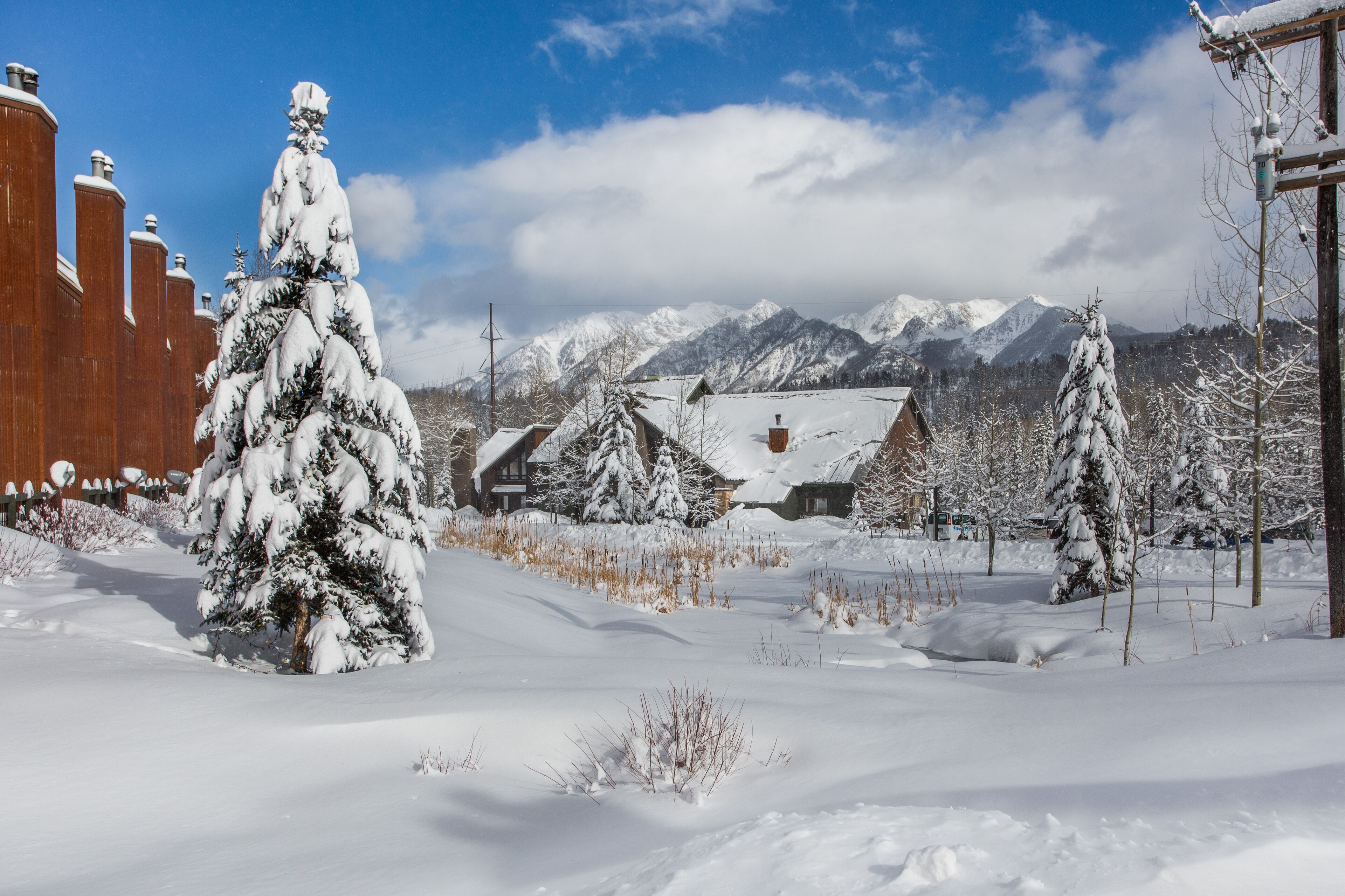 A snow-covered landscape with a wooden building, snow-covered trees, and mountains in the background.