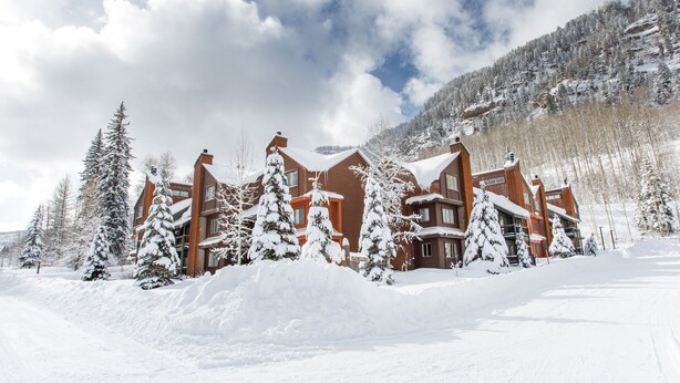 Un gran albergue de varios pisos con techo nevado y árboles circundantes, con montañas al fondo.