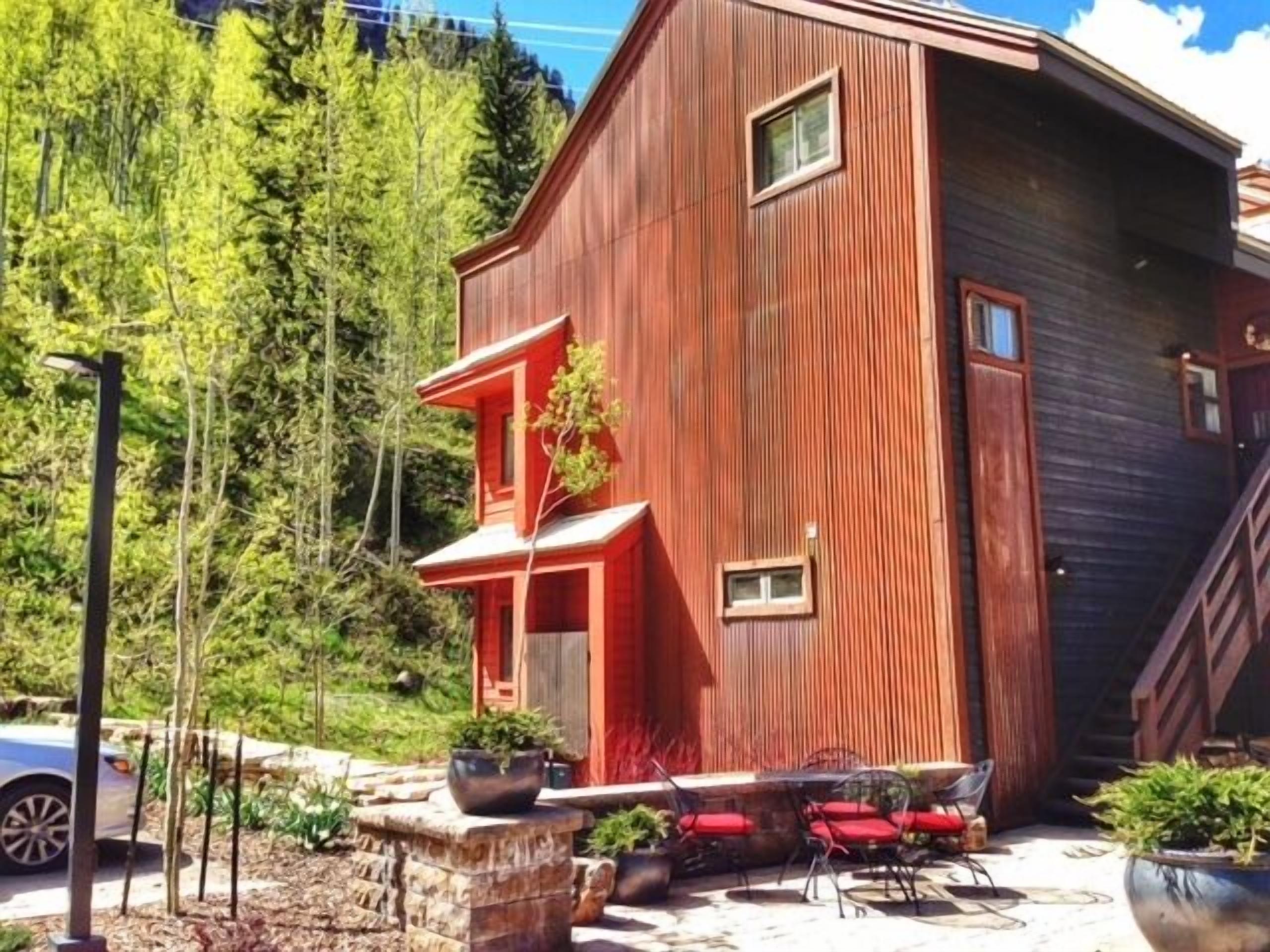 A modern wooden house with a unique architectural design, featuring a sloped roof and a small porch area with outdoor seating.