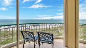 A balcony with two chairs overlooking a beach with umbrellas and people.