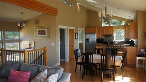 Amplia sala de estar con sofá, mesa de comedor y sillas. Se ve una cocina con refrigerador y alacenas al fondo.