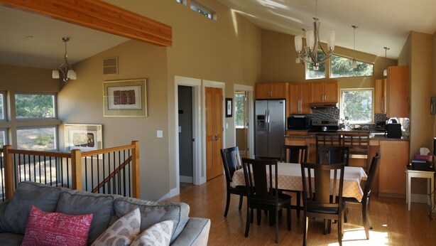 A spacious living room with a sofa, dining table, and chairs. A kitchen with a refrigerator and cabinets is visible in the background. - Luxurious Home W/ Bay & Ocean Views - 2 of 3 Bdrms Masters! (Bay City)