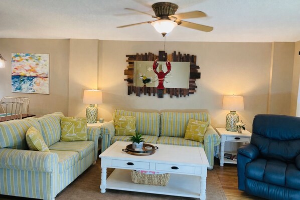 A living room with striped sofas, a coffee table, a ceiling fan, and a wall-mounted abstract art piece. - Ocean Village Club R17 Exceptionally Clean! On the Beach! OVER 280 "5 STARS" (St. Augustine)