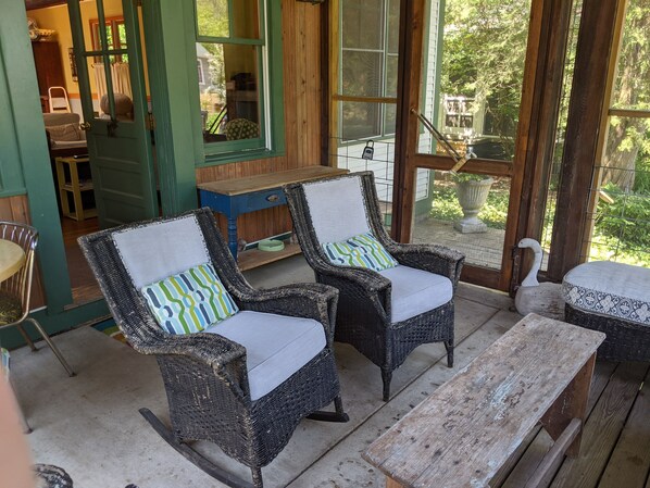 A porch with wicker rocking chairs, a wooden table, and a view of a house through a screened door.
