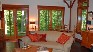 A cozy living room with a sofa, wooden coffee table, and a lamp. There are large windows with a view of trees. - Timber Ridge Cabin Apartment, Forested Retreat Close to Natchez Trace, Franklin! (Franklin)