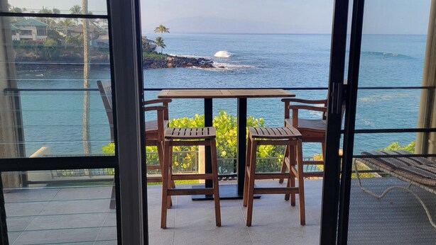 A balcony with a table and two chairs, overlooking a coastal view with waves and a distant island. - Ocean Front Napili Point C20 Amazing View  A/C  Freshly Remodeled for 2025! (Napili, Lahaina)