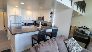 A modern kitchen with white cabinets, a stainless steel refrigerator, and a granite countertop. There is a dining area with a gray sofa and patterned cushions. - Ocean Front Napili Point C20 Amazing View  A/C  Freshly Remodeled for 2025! (Napili, Lahaina)