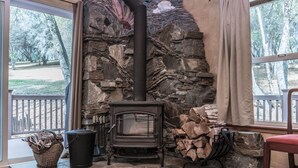 Ein Steinkamin mit schwarzem Schornstein, daneben gestapelte Holzscheite und ein Fenster mit Blick auf Bäume.