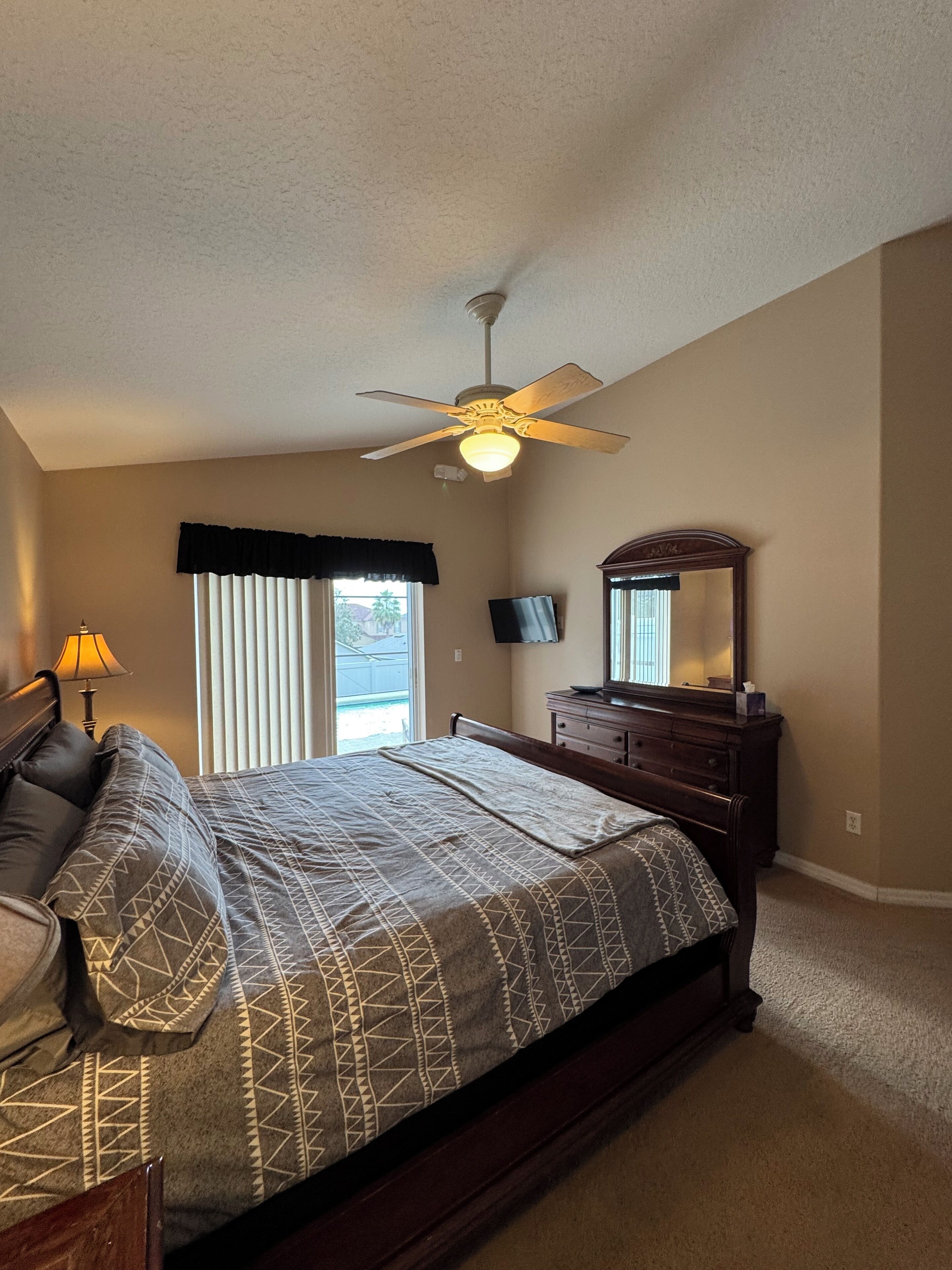 A bedroom with a bed, a dresser, a ceiling fan, a TV, and a window with blinds.