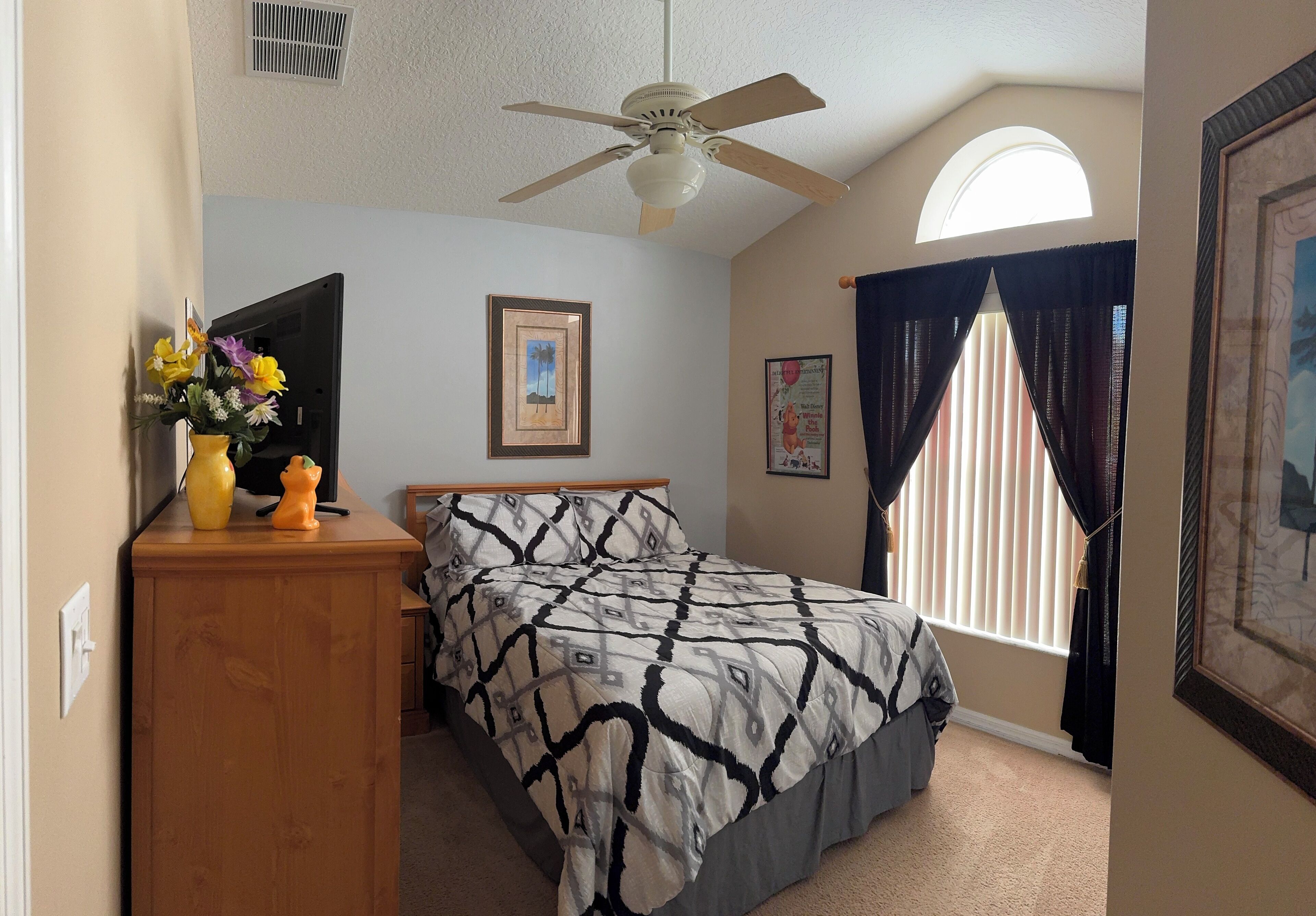 A bedroom with a bed, a ceiling fan, a dresser, a window with curtains, and framed pictures on the walls.