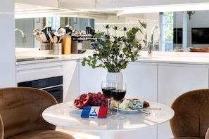 A modern kitchen with a dining area, featuring a table with a glass vase, pomegranate, grapes, cheese, and wine glasses. - Spacious, Enchanting 1 Bedroom + Balcony and Insta-worthy Eiffel View (Paris)