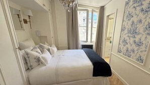 A neatly made bed with white and blue bedding, a chandelier, and a window with curtains. - Spacious, Enchanting 1 Bedroom + Balcony and Insta-worthy Eiffel View (Paris)