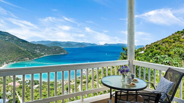 A balcony with a table and chairs, overlooking a turquoise sea and green hills.