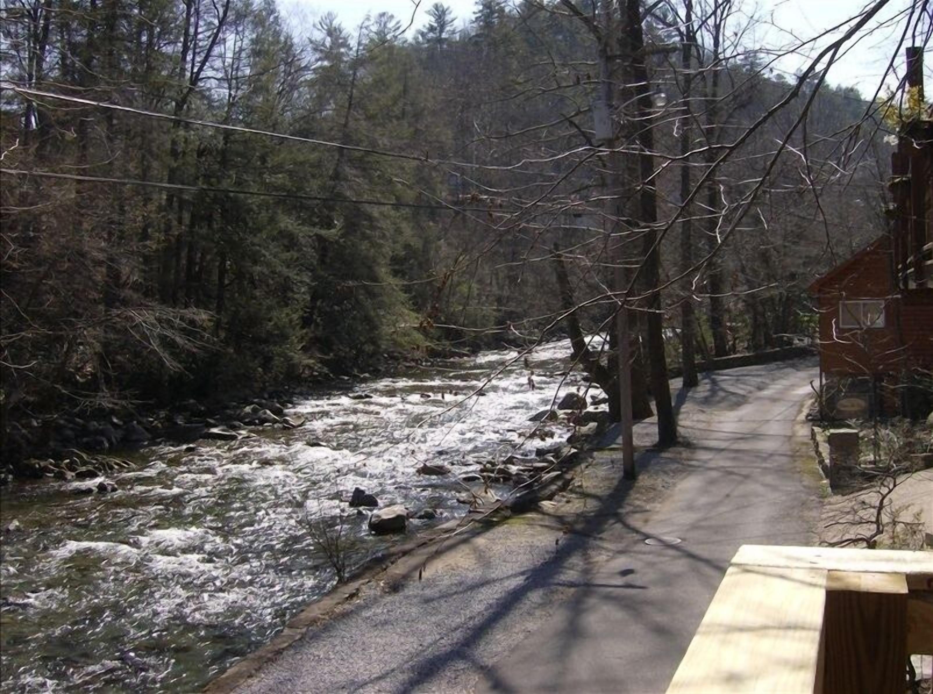 A wooden bench by a river with a road and a house in the background.