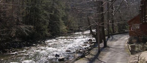 A wooden bench by a river with a road and a house in the background.