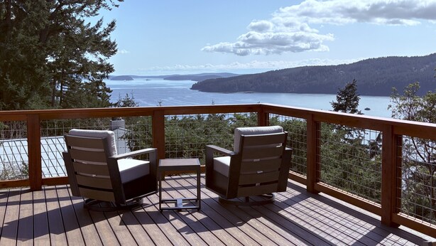 A wooden deck with two chairs and a small table, overlooking a lake and forested hills. - Orcas-Beautiful Modern Home- Large New Deck- Water View- Privacy- Dog Friendly (Rosario)