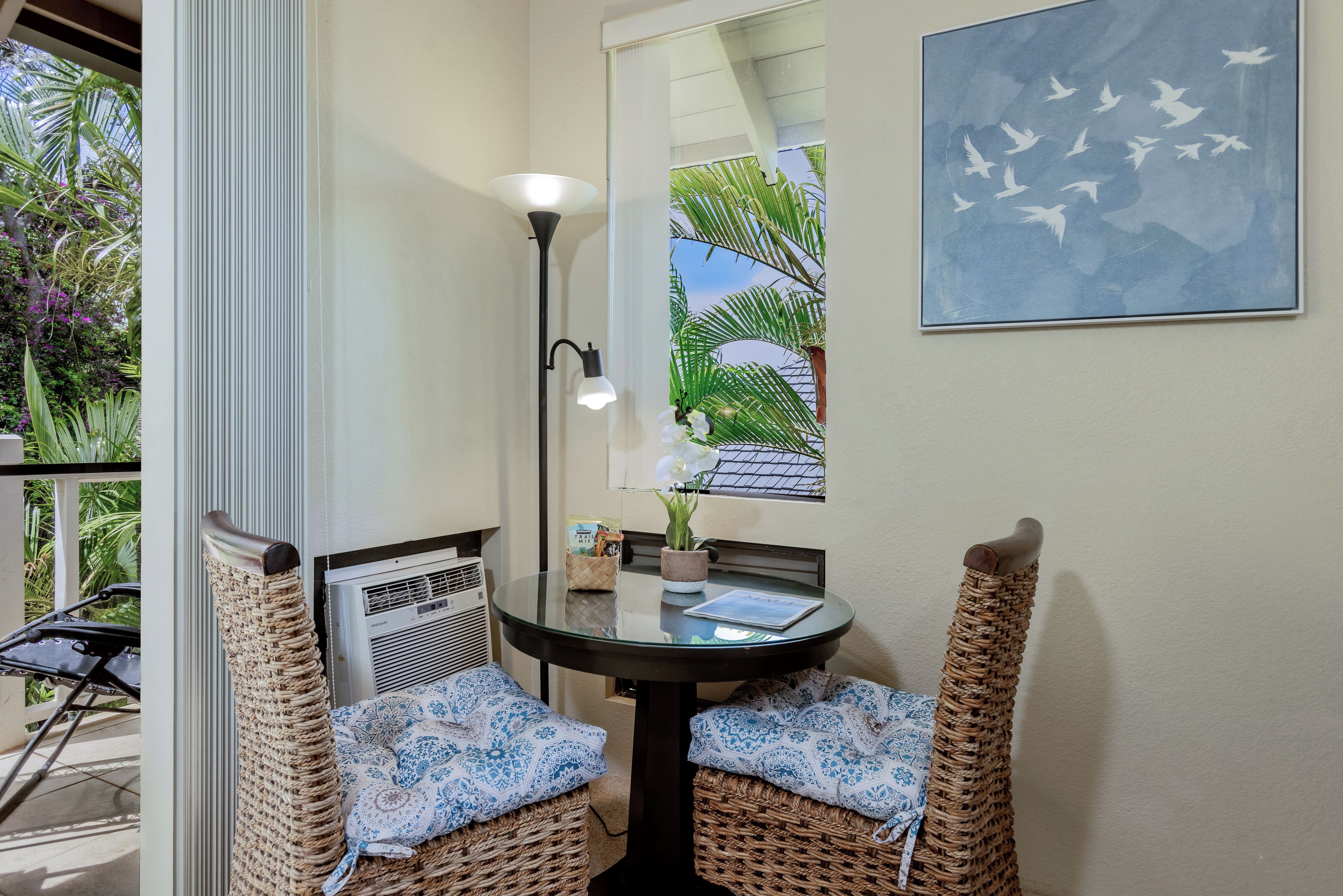 A room with a round glass table, two wicker chairs with cushions, a floor lamp, a wall-mounted mirror, and a framed picture.